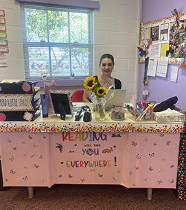 Mrs. Labra sitting at her desk which is cover in pink paper and butterflies. The sign says Reading will take you everywhere.